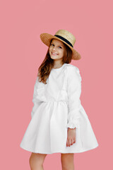 Front image of a cheerful little girl in cute white dress, straw hat, looking at camera, on a pink background.