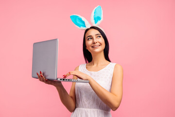Thoughtful woman in bunny ears holding laptop, video call, shopping online in studio on pink background, easter