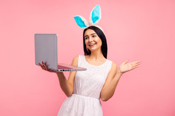 Positive woman in rabbit ears holding laptop, video call, shopping online in studio on pink...