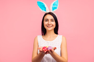 Cute brunette woman in bunny ears holds painted Easter eggs in her hands, Easter, in the studio on a pink background