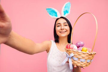 beautiful cheerful woman in rabbit ears takes a selfie, holds a basket with yayyuami in her hands, video call, easter, pink background