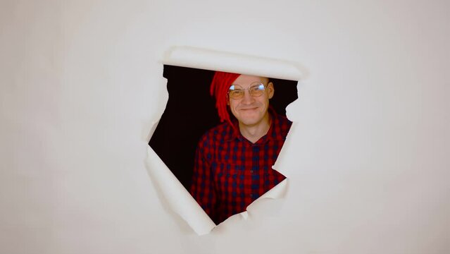 Young man grimacing face in studio. Crazy guy in glasses aping and sticking out of hole of white background.