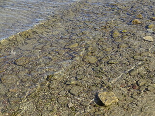 Close up of Baltic sea ripple on water surface with sunlight reflecting on a sunny day. Various size smooth brown stones, pebble under water