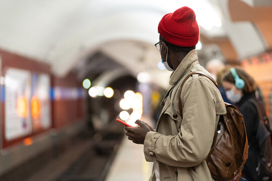 African American hipster man in red hat wear mask in subway using smartphone waiting for train on platform. Black student male with mobile phone at underground. Covid-19, transportation concept.