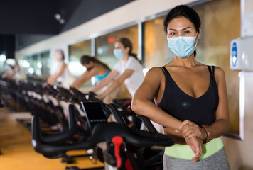 Portrait of young adult woman in protective face mask posing near stationary bike at gym