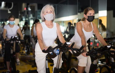 Fototapeta na wymiar Portrait of young and senior women wearing face mask for disease protection training together during stationary bike workout in gym