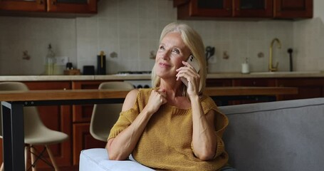 Smiling middle-aged woman make call enjoy pleasant personal lively conversation on smartphone sits on sofa at home, spend leisure speaks to friend use cellphone. Mobile carrier operator, tech concept