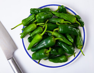 Plate full of fresh green padron peppers and steel kitchen knife on white background. Organic cooking ingredients..