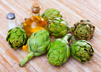 Pile of fresh artichokes on wooden table with olive oil in glass decanter and seasonings. Organic...