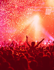 Naklejka na ściany i meble A crowded concert hall with scene stage in red lights, rock show performance, with people silhouette, colourful confetti explosion fired on dance floor air during a concert festival