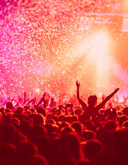 Naklejka na ściany i meble A crowded concert hall with scene stage in red lights, rock show performance, with people silhouette, colourful confetti explosion fired on dance floor air during a concert festival