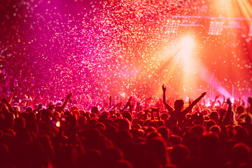 A crowded concert hall with scene stage in red lights, rock show performance, with people silhouette, colourful confetti explosion fired on dance floor air during a concert festival - obrazy, fototapety, plakaty