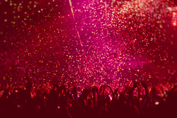 A crowded concert hall with scene stage in red lights, rock show performance, with people silhouette, colourful confetti explosion fired on dance floor air during a concert festival