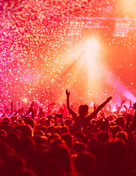 A crowded concert hall with scene stage in red lights, rock show performance, with people silhouette, colourful confetti explosion fired on dance floor air during a concert festival