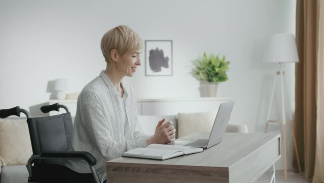 Virtual business meeting. Happy middle aged woman wheelchair user waving hand to laptop, greeting her partners online