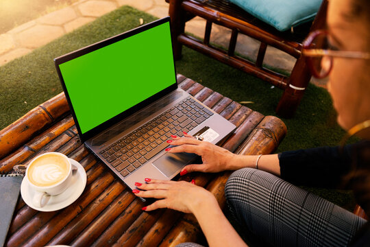 Latin business woman with curly hair and glasses checks her computer with green screen in a restaurant with a cappuccino coffee next to her. Digital entrepreneurship concept.
