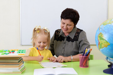 Mature teacher sitting at table teaching little girl how to write