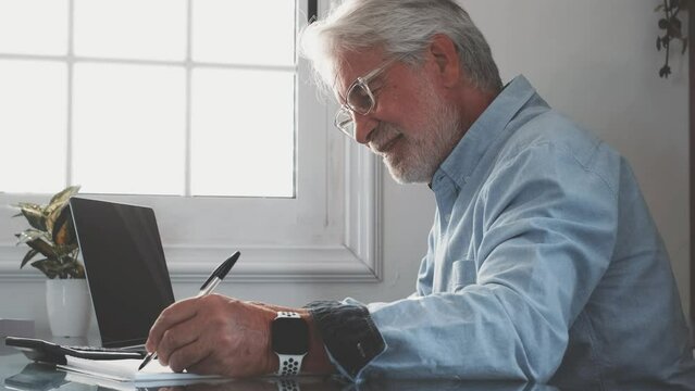 Happy old caucasian businessman smiling working online watching webinar podcast on laptop and learning education course conference calling make notes sit at work desk, elearning concept
