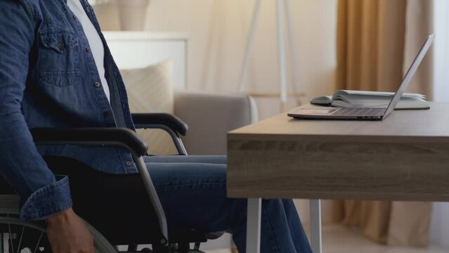 Freelance for everyone. Unrecognizable man wheelchair user riding to table and start typing on laptop at home