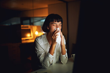 Risking her health for the sake of the deadline. Shot of a young businesswoman blowing her nose during a late night at work.