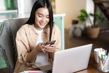 Asian female student looking at mobile phone, sitting at home with laptop, register online course...