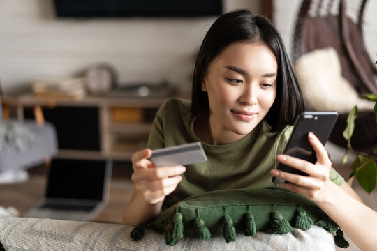 Young asian woman buying from online shop, using mobile phone and credit card, shopping from home