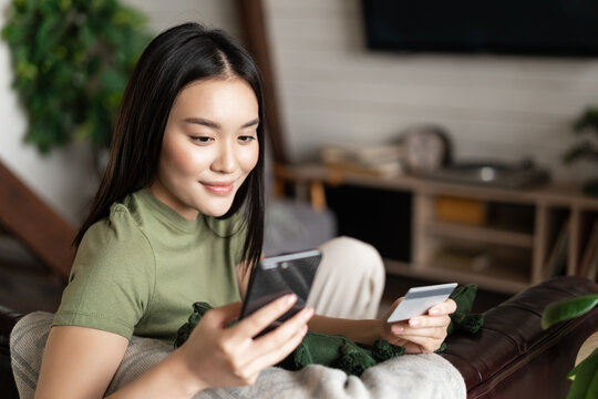 Smiling Korean Girl Using Mobile Phone And Credit Card While Sitting At Home, Paying For Purchase In Internet. Buying Something From Her House