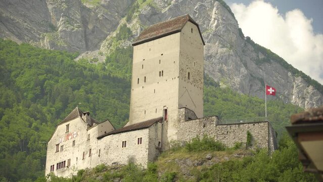 Low angle of historical Sargans Castle with Swiss flag waving on a hill near Mount Gonzen surrounded by dense green pine forest, Switzerland Alps