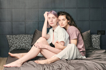 Portrait of serious young Caucasian women in sleepwear sitting on satin linen and hugging in bedroom