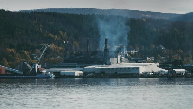 A working ferrosilicon production plant on the shore of the fjord. Smoke and steam are rising from the chimneys. Crane unloading goods. Heavy machinery moves on the site.