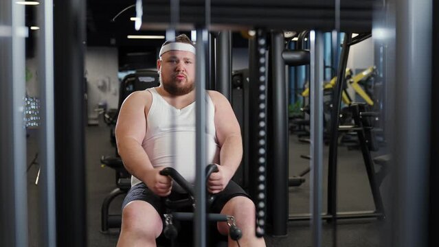 Fat burning, overweight funny man lifting blocks on rowing machine, training on block device and gym equipment, motivation.