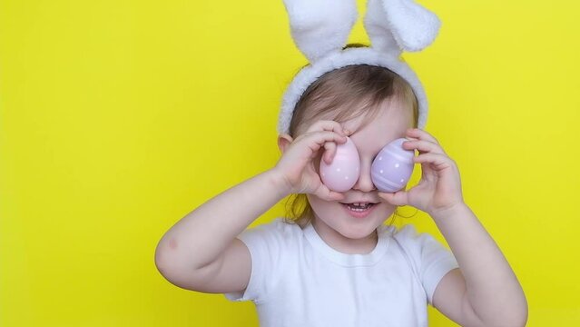 Little girl in rabbit bunny ears on head looking into the camera with colored Easter eggs on yellow background.