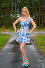 Beautiful caucasian woman in a mini blue-white dress and high heel sandals standing on the wet road in a city park in Tallinn on a cloudy summer day and looking into the camera.