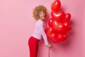 Happy curly haired young European woman wears jumper and red trousers stands sideways holds helium heart balloons has leaked makeup romantic mood feels energetic poses against pink background