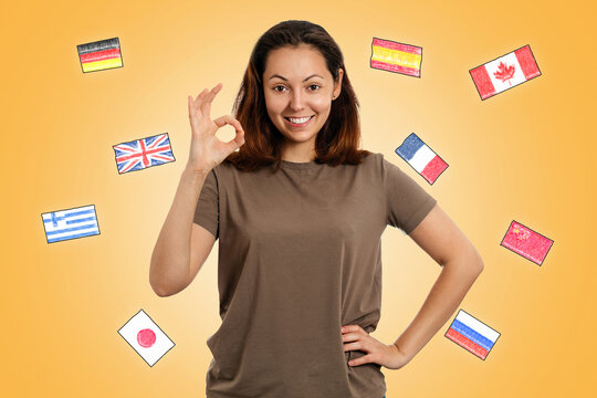 English language Day. A young smiling woman makes an OK gesture. Yellow background with flags of different countries. The concept of learning foreign languages