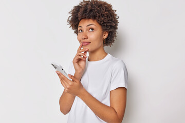 Sideways shot of curly haired young woman has dreamy expression has thoughtful expression uses smartphone thinks what answer to give dressed casually isolated over white bckground. Technology