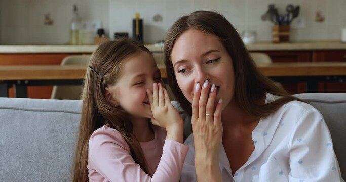 Little adorable daughter share secrets on mothers ear, cover mouth telling confidential information gossiping together seated on cozy sofa at home. Trust and understanding between mom and kid concept