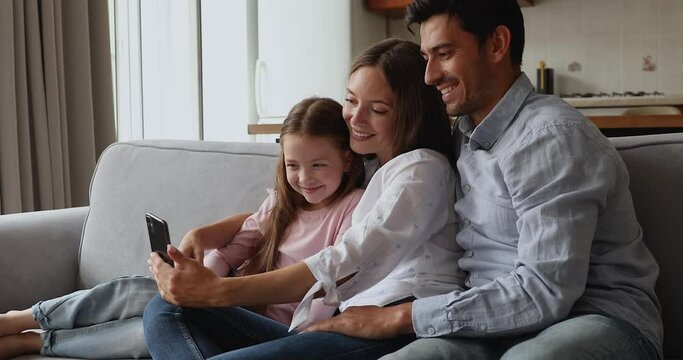 Young married couple and cute daughter relax on sofa look at smartphone screen make selfie pictures for memory, use new mobile app spend funny leisure at home. Modern tech, e-commerce clients, concept