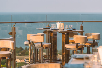 Stylish table and stools of an outdoor restaurant on the rooftop of a building, utensils are set on the table, glass fence, blurred background of sky and sea, evening sunlight, no people.