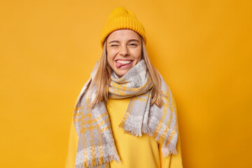 Positive carefree young woman winks eye and sticks out tongue wears hat casual jumper scarf around...