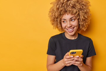 Fototapeta na wymiar Positive good looking young woman with cheerful smile on face curly hair uses smartphone chats online wears casual black t shirt isolated over yellow background blank space for your advertising