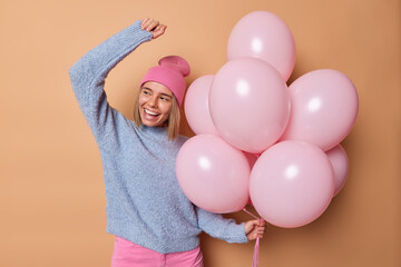 Horizontal shot of positive European woman wears hat casual blue jumper dances carefree enjoys birthday celebration holds bunch of inflated balloons isolated over brown background. Festive occasion