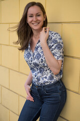 Portrait of smiling brunette woman standing in front of yellow wall, wearing printed blouse and jeans