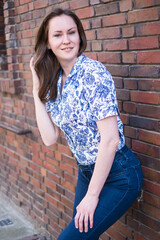 Portrait of brunette smiling woman standing in front of brick wall and stroking hair, wearing printed blouse and jeans