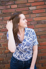Portrait of brunette smiling woman standing in front of brick wall and stoking her hair, wearing printed blouse and jeans