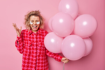 Fototapeta na wymiar Angry irritated woman shouts fromm anger keeps hand raised holds bunch of inflated balloons prepares for holiday party applies beauty patches under eyes dressed in checkered jacket poses indoor