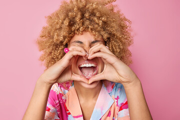 Optimistic glad woman with curly bushy hair makes heart gesture over mouth says I love you shows...