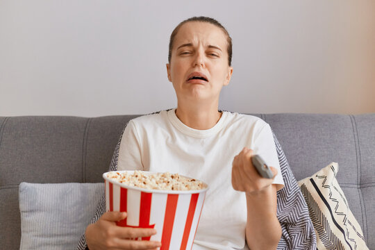 Indoor shot of sad upset young woman wearing white t shirt sitting on sofa with popcorn and remote control, watching romantic movie or sees tragic moment in the serial.