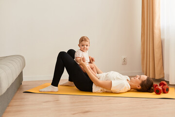 Image of adorable woman wearing white t shirt and black leggins doing sport exercises with baby daughter, having fun with her kid while having workout.