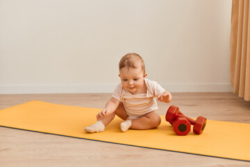 Horizontal shot of charming cute toddler baby sitting on yoga mat with dumbbells, exploring sport equipment, wearing bodysuit.
